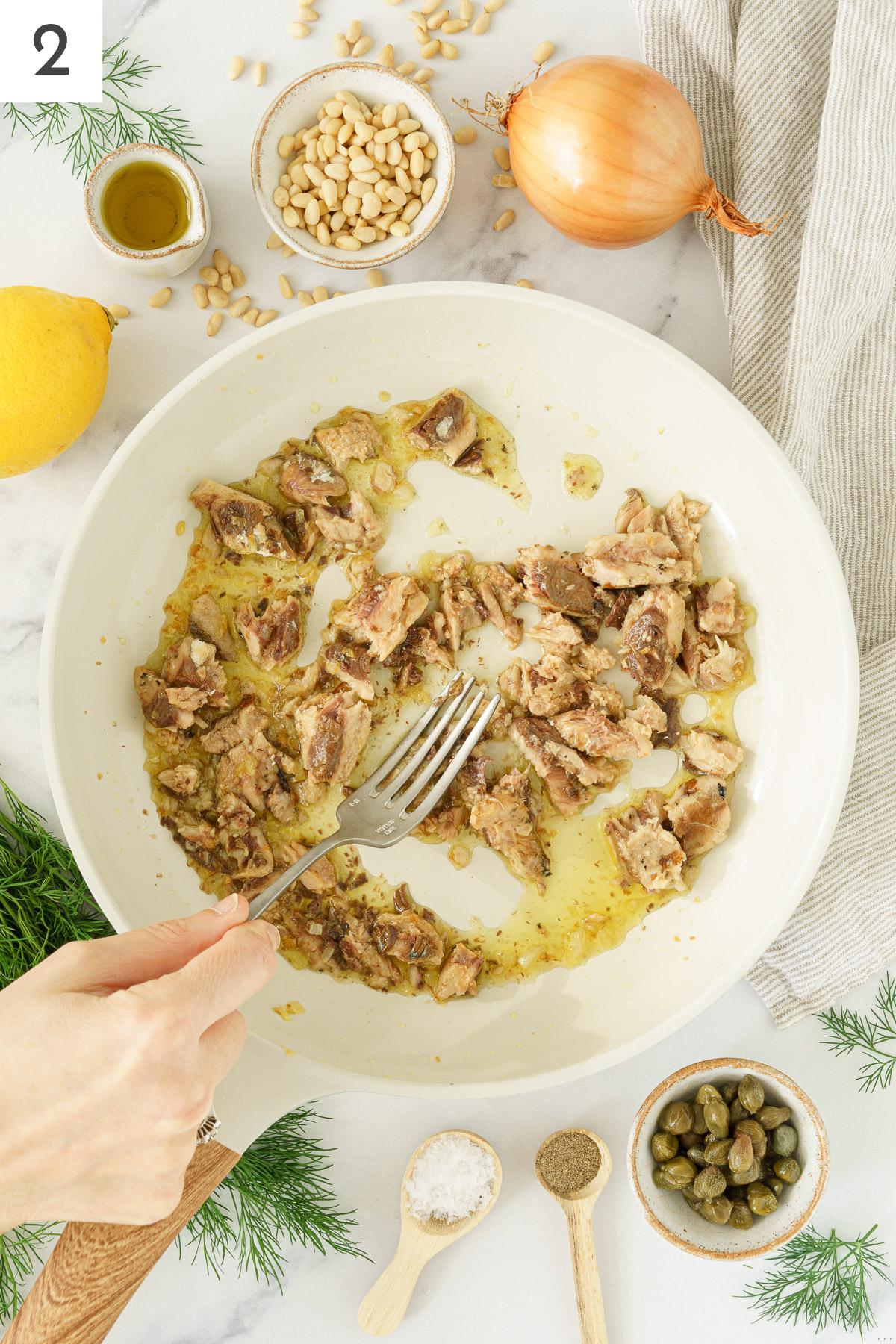 A hand gently breaking the sardines in a pan with sauteed onions.