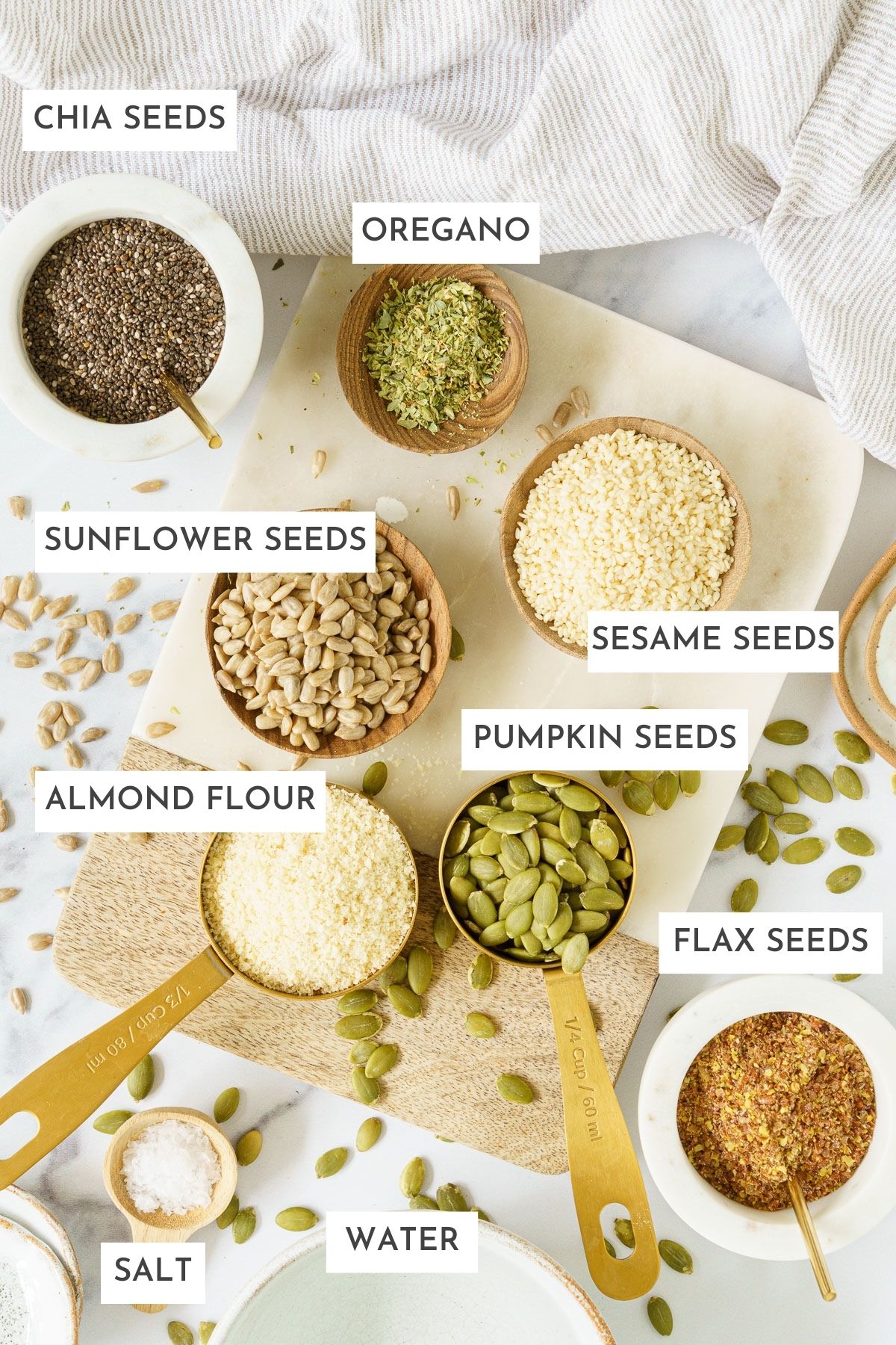 Laid out ingredients of seed crackers: Oregano, chia seeds, sunflower seeds, sesame seeds, pumpkin seeds, almond flour, flax seeds, water, and salt.